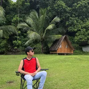 Joowd sitting outdoors on a camping chair, cap and sunglasses on, palm trees behind